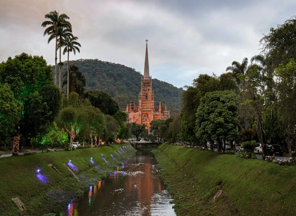 View of the Cathedral of Saint Peter of Alcantara in Petrópolis, Rio de Janeiro, Brazil, surrounded by trees and mountains, reflecting on the Piabanha River at dusk.