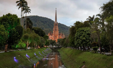 View of the Cathedral of Saint Peter of Alcantara in Petrópolis, Rio de Janeiro, Brazil, surrounded by trees and mountains, reflecting on the Piabanha River at dusk.
